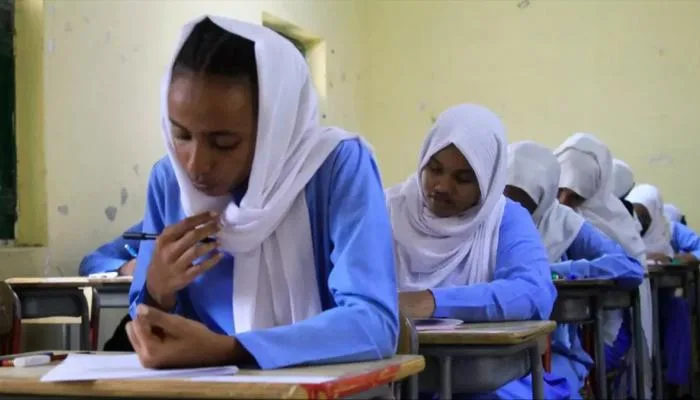 طلاب وافدون أمام مبنى جامعي في السودان يطّلعون على أوراق التقديم، مع إشارة إلى موعد التقديم للعام الدراسي 2026–2027 ورسوم 300 دولار.